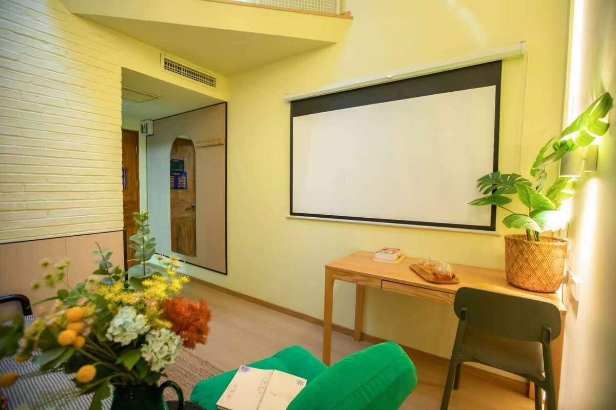 Une pièce avec une grande fenêtre, un bureau en bois, une chaise verte, une petite table avec une plante et un tableau blanc accroché au mur.