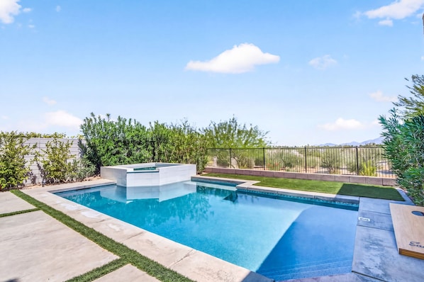 Piscina rectangular con jacuzzi integrado, rodeada por un jardín ajardinado con arbustos variados y una cerca.