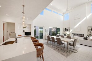 A modern kitchen with a central island, dining area with chairs, and a living space with a sofa and artwork. - Palladium Palace: Pool+Spa, EV Charger, Miralon! (Palm Springs)