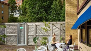 An outdoor seating area with white plastic chairs and tables, a wooden fence with a gate, and a building with a blue awning. - 1 Person Holiday Park Home in Goteborg (Gothenburg)
