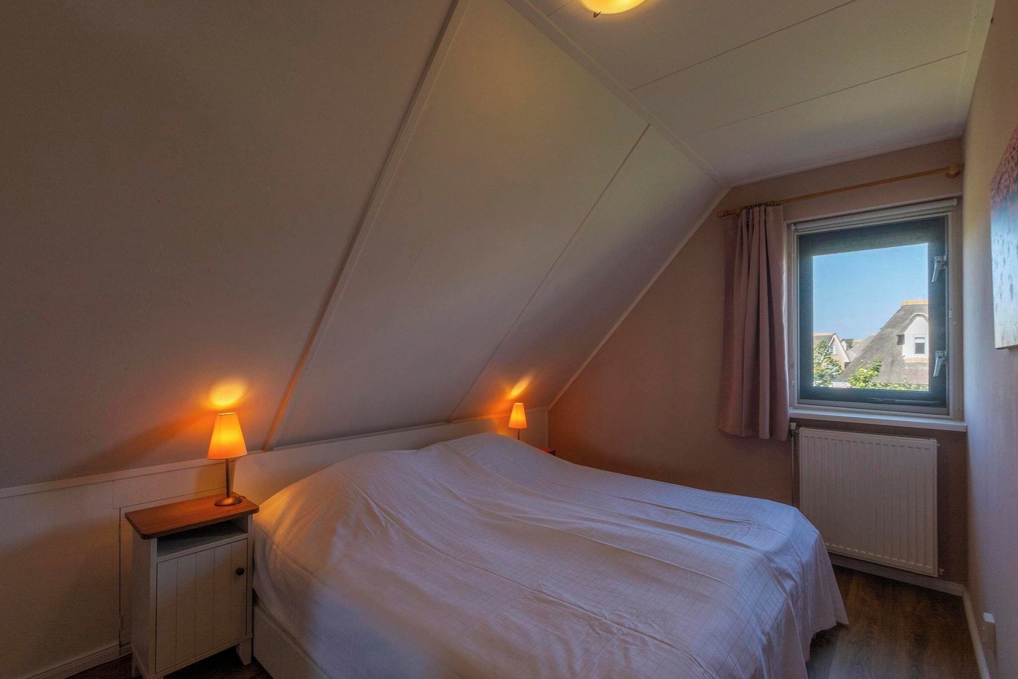 Een kleine kamer met een enkel bed, schuine kap, raam met gordijnen en twee bedlampjes.