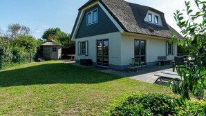 Una casa de dos pisos con techo oscuro y paredes claras, un patio con mobiliario exterior y un pequeño cobertizo en el jardín trasero.