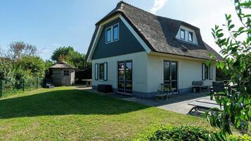 Een tweelaags huis met een donker dak en lichte muren, een terras met tuinmeubilair en een kleine schuur in de achtertuin.