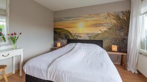 Una camera da letto con un letto, una scrivania, una sedia, un comodino, una lampada e un quadro raffigurante un tramonto sulla parete.