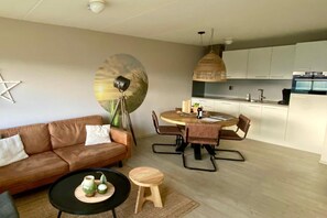 Ett modernt vardagsrum med en brun lädersoffa, ett runt matbord med stolar, en köksdel med vita skåp och en hängande lampa.