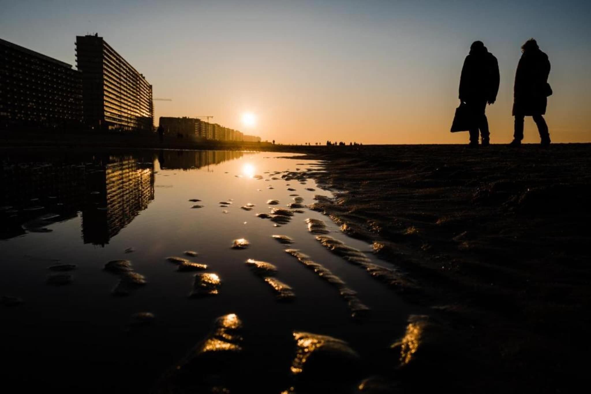La silhouette de deux personnes se promenant au bord de l’eau au coucher du soleil, avec des bâtiments en arrière-plan.
