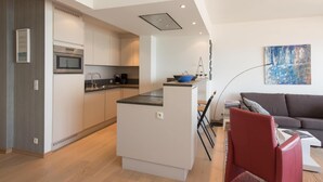 Una cocina moderna con armarios blancos, horno empotrado y encimera negra. Hay un comedor con una silla roja y una mesa blanca. Un cuadro adorna la pared, y un sofá se encuentra en la sala de estar.