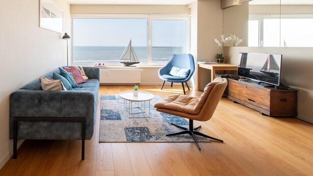Een moderne woonkamer met een bank, fauteuil, salontafel en televisie. Een model zeilboot staat op de vensterbank, en er staat een plant op de salontafel.