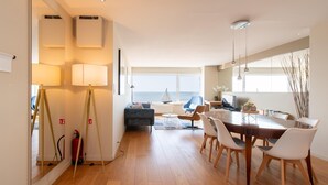 Een ruime woonkamer met houten vloer, een eettafel met stoelen, een bank en uitzicht op zee door grote ramen.
