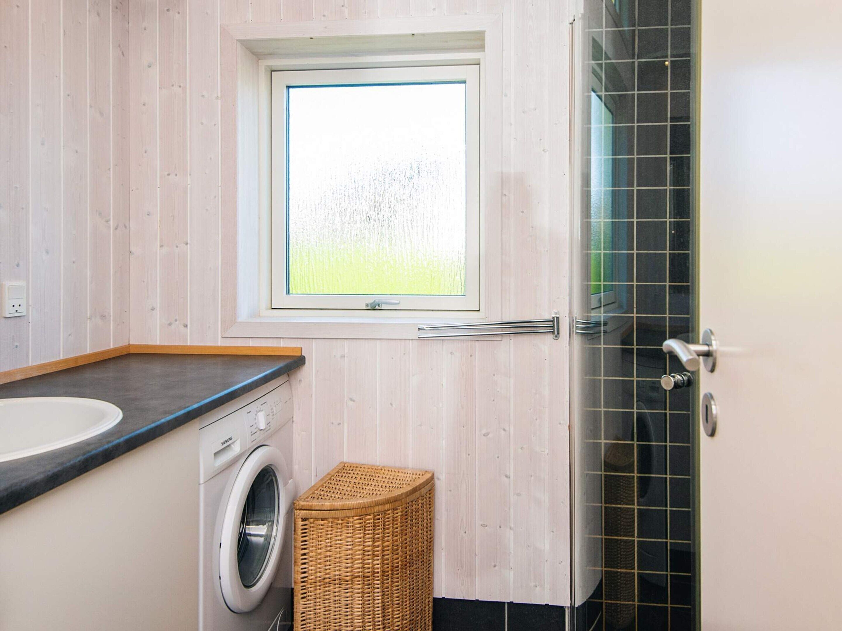 Un baño pequeño con lavadora, una cesta de mimbre y ventana con vistas a la vegetación.