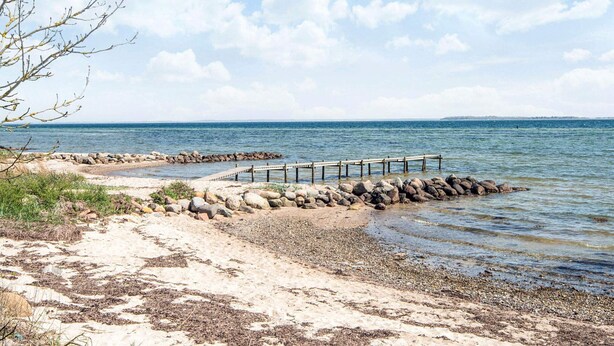 A coastal scene with a wooden pier extending into calm waters, surrounded by a rocky shoreline and sparse vegetation. - 4 Star Holiday Home in Hejls (Hejls)