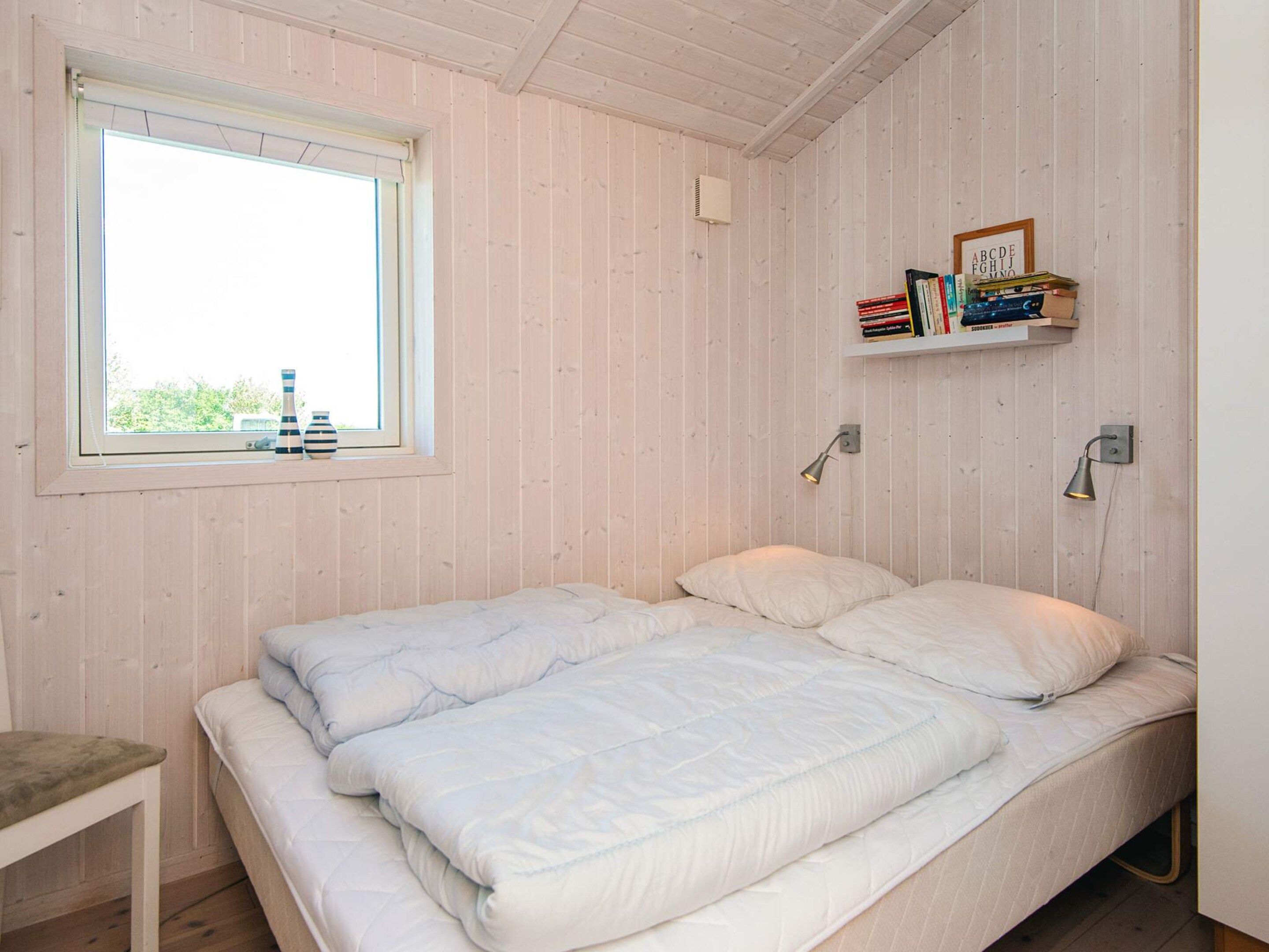 A bedroom with a single bed, a window, a bedside table, and a shelf with books.