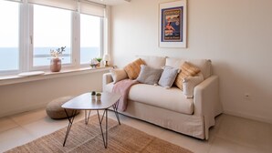 A modern living room with a white sofa, a small coffee table, and a window with a view of the sea. - Apartment in Ostend with Stunning Sea View (OSTEND)