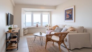 A modern living room with a white sofa, a wicker chair, a small table, and a TV on a shelf. - Apartment in Ostend with Stunning Sea View (OSTEND)