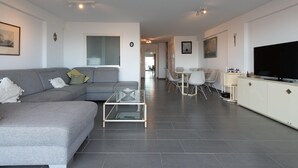 Una sala de estar moderna con un sofá gris, una mesa de centro de vidrio, una zona de comedor con sillas y un televisor sobre un mueble.