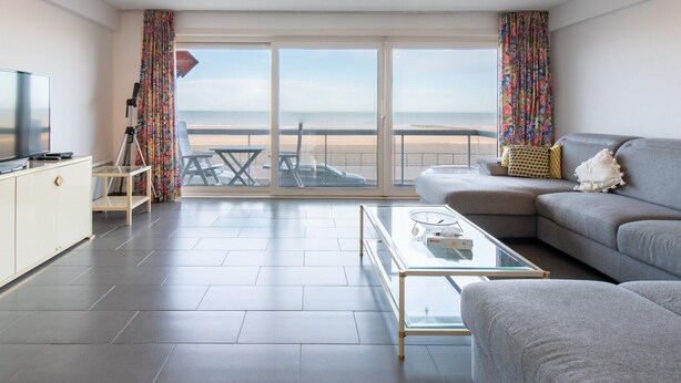 Una sala de estar moderna con un sofá, una mesa de centro de vidrio, un televisor y un balcón con vista a la playa.