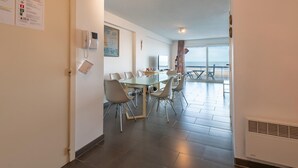 Una habitación de hotel moderna con zona de comedor que incluye una mesa de vidrio y sillas, un televisor de pantalla plana y vistas al mar a través de amplios ventanales.