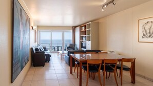 A modern living room with a dining area, a sofa set, and a large window offering a view of the sea. - Apartment in Mariakerke with Stunning Sea Views (OSTEND)