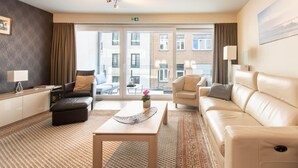 A modern hotel room with a large window, a sofa, a chair, a coffee table, and a small table with a lamp. - Apartment by the Beach in Ostend with Terrace (OSTEND)