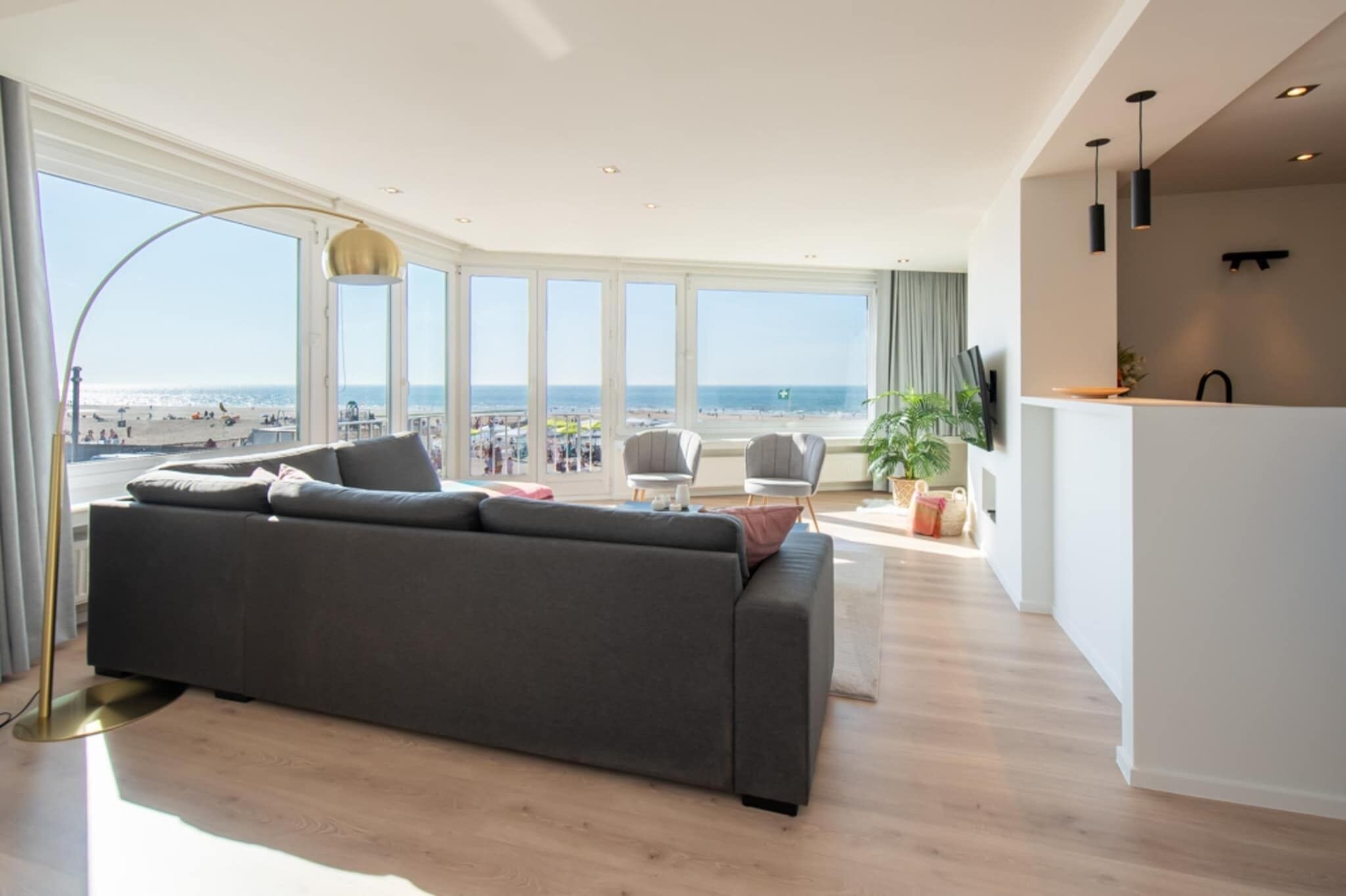 Een moderne woonkamer met een grote grijze bank, een vloerlamp en uitzicht op het strand.