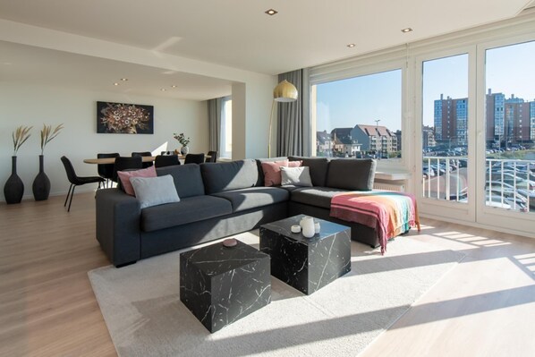 A modern living room with a sectional sofa, a dining area, and large windows offering a city view. - Apartment in Ostend with Stunning Sea Views (OSTEND)