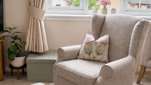 A living room with a beige armchair, a matching ottoman, a small side table with a plant, and a window with curtains.