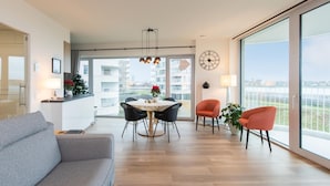 A modern living room with a grey sofa, a dining table with chairs, and a large window offering a city view.
