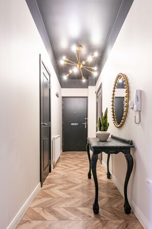 Un corridoio con pavimento a spina di pesce, una porta nera, uno specchio con cornice decorativa, una pianta in vaso e un telefono fissato al muro.
