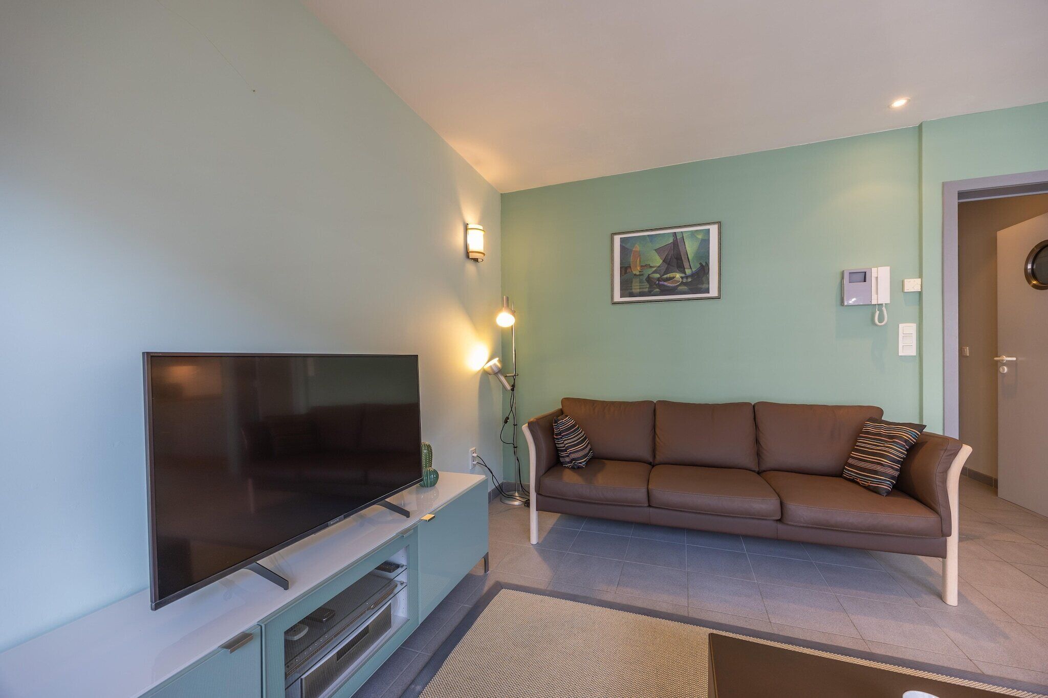 A living room with a flat-screen TV, a brown leather sofa, a painting on the wall, and a door in the background.