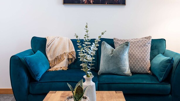 Un canapé de couleur turquoise agrémenté de coussins à motifs, une table basse en bois sur laquelle est posé un vase décoratif, et une œuvre d’art encadrée au-dessus.