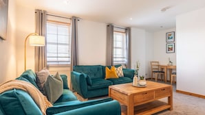 Un salon avec un canapé turquoise, une table basse en bois et un lampadaire. Il y a deux fenêtres équipées de stores, un coin repas avec une table et des chaises en bois, et un mur décoré de tableaux encadrés.
