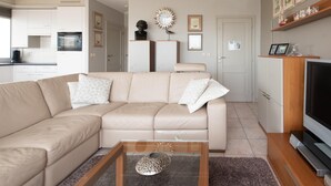 Een moderne woonkamer met een beige modulaire bank, een glazen salontafel en een flatscreen televisie. Er is een keukenhoek met witte kasten en een magnetron.