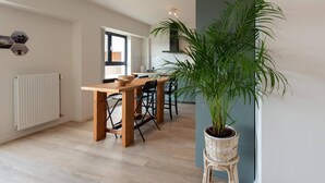 A modern kitchen with a dining area, featuring a wooden table and chairs, a large potted plant, and a view into the adjacent dining room. - Apartment in Ostend near the Beach (OSTEND)