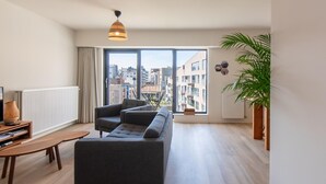 A modern living room with a grey sectional sofa, a wooden coffee table, and a large window offering a city view. - Apartment in Ostend near the Beach (OSTEND)