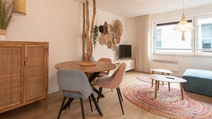A modern living room with a dining area, featuring a round wooden table, two chairs, a sofa, a TV, and a large window with natural light. - Apartment in Ostend near the Beach (OSTEND)