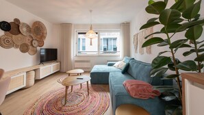 A modern living room with a sofa, a coffee table, a TV on a stand, a large plant, and a colorful rug. - Apartment in Ostend near the Beach (OSTEND)