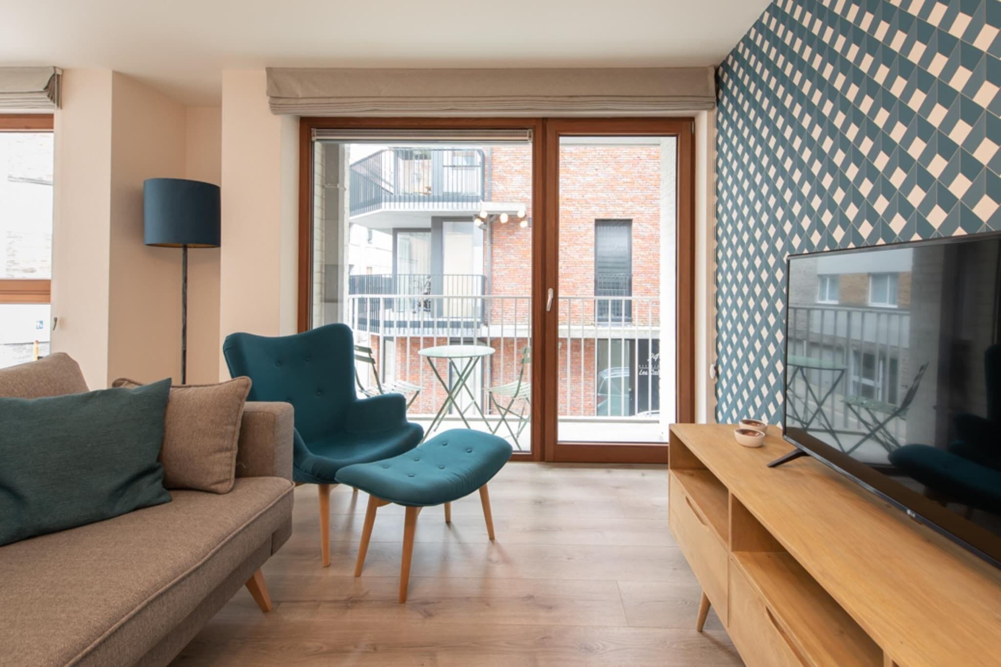 A modern living room with a sofa, teal armchair, and ottoman, a wooden TV stand, and a large TV.