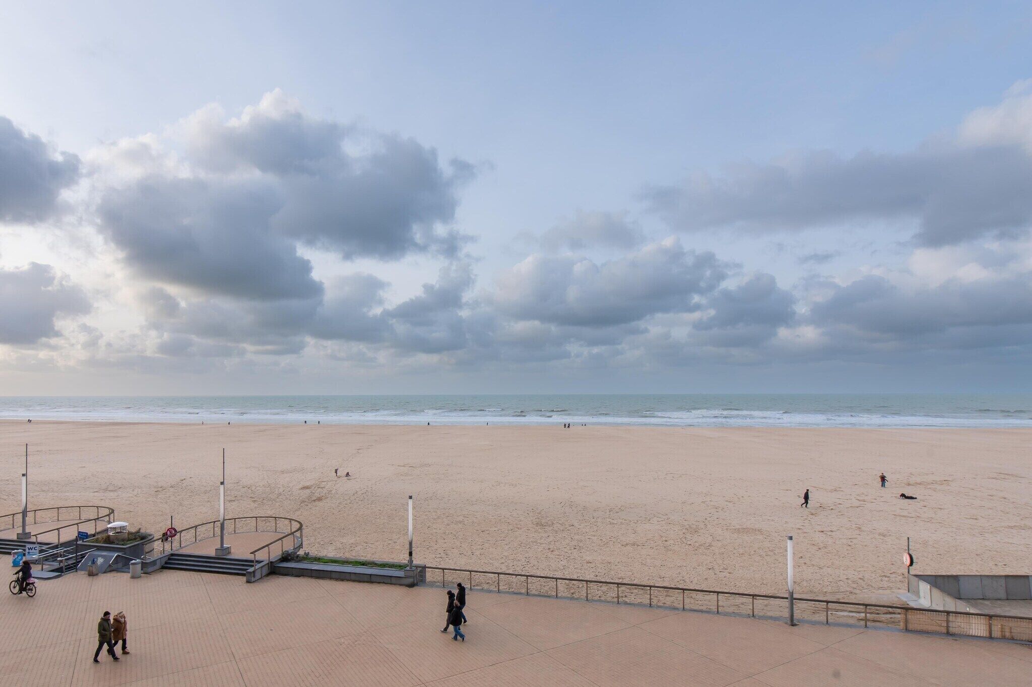 Una playa con un paseo marítimo pavimentado, algunas personas caminando y un cielo nublado.