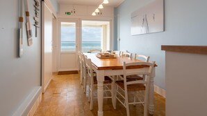 A dining area with a wooden table and white chairs, a view of the ocean through a glass door, and a framed artwork on the wall.