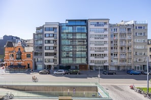 A multi-story building with a modern glass facade, balconies, and a parking area with cars and bicycles.
