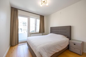 Ein Schlafzimmer mit Bett, Nachttisch, Fenster mit Vorhängen und Blick auf Gebäude.
