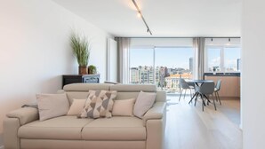 A modern living room with a beige sofa, a dining area with a table and chairs, and a large window offering a city view. - Apartment in Ostend near Marina & Park (OSTEND)
