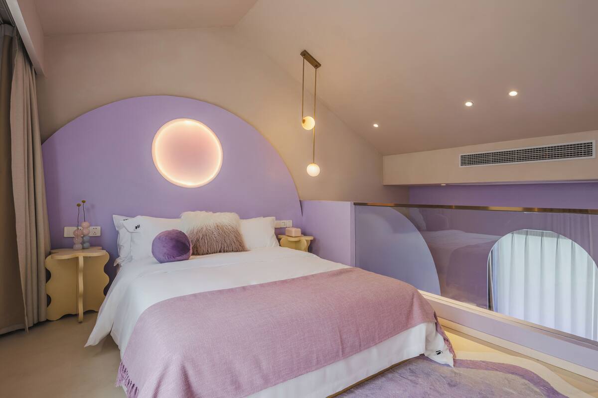 A modern bedroom with a curved ceiling, a large bed with a pink and white bedspread, a round wall light, and a bedside table with a vase.