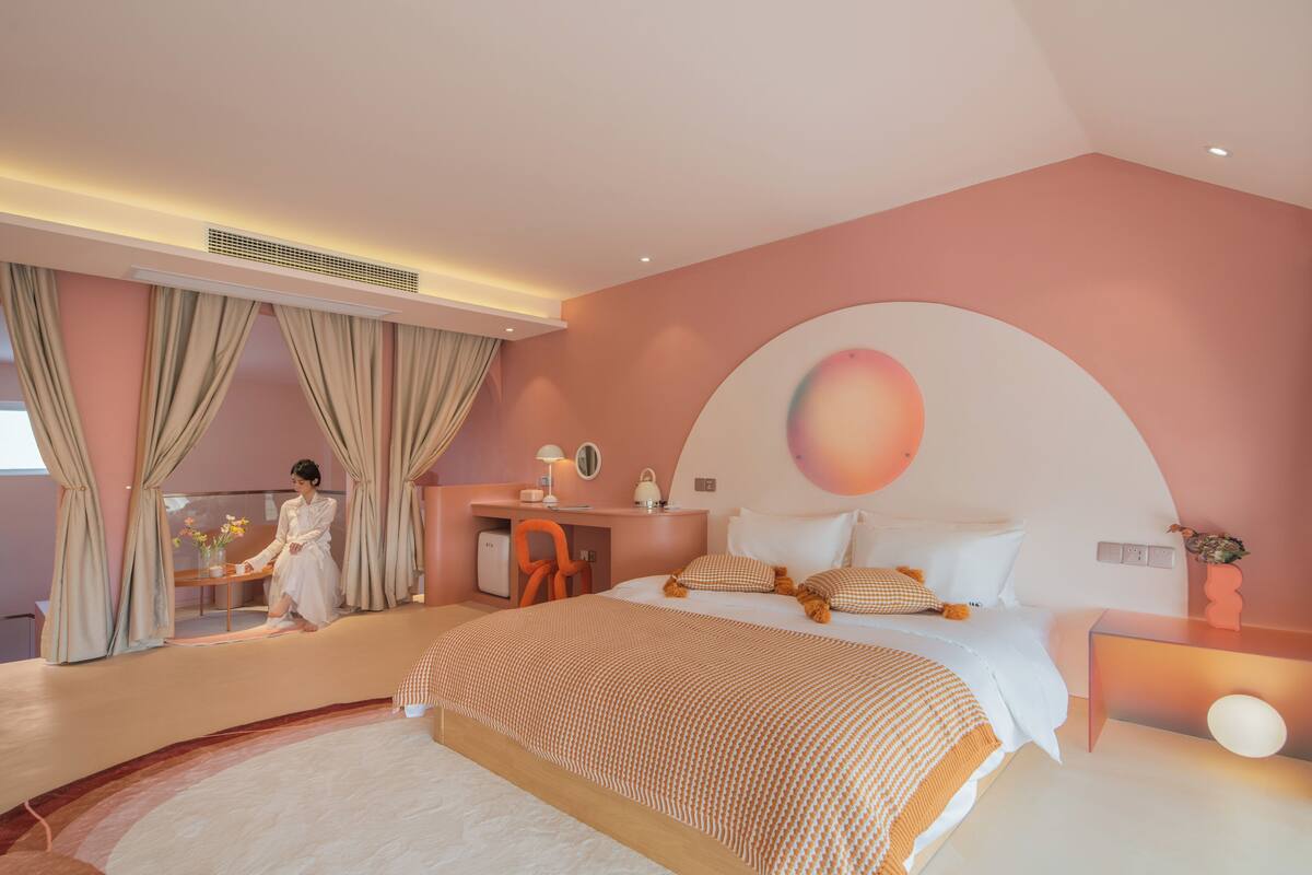A modern bedroom with a curved ceiling, a large bed with patterned bedding, a desk with a chair, and a large window with curtains.