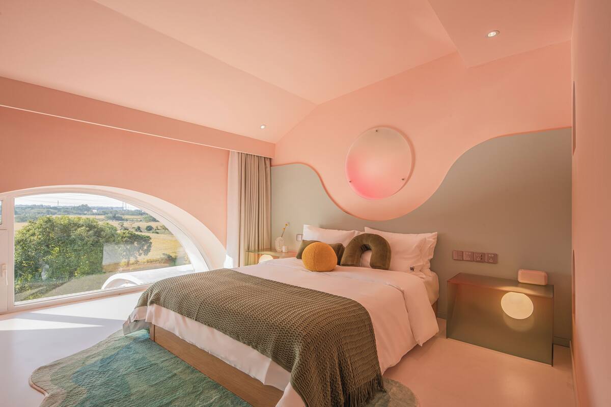 A bedroom with a large window, a bed with a green blanket, a round mirror, and a bedside table with a lamp.