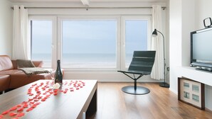 Een moderne woonkamer met een houten vloer, een bank, een eettafel versierd met rozenblaadjes, een stoel, een televisie en uitzicht op zee door grote ramen.
