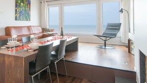 Een eetgedeelte met een houten tafel gedekt voor vier personen, een bank en een stoel bij een groot raam met uitzicht op zee.