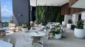 A modern lounge area with a view of the ocean, featuring a sofa set, dining tables, and potted plants. - La Jolla Holiday Broadbeach (Broadbeach)