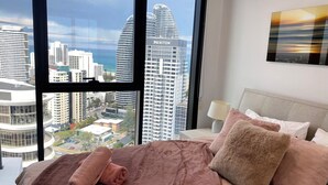 A hotel room with a large window offering a city view, a bed with a pink blanket, and a nightstand with a lamp. - La Jolla Holiday Broadbeach (Broadbeach)