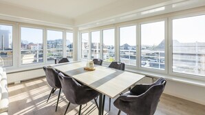A modern dining room with a large table and chairs, large windows with a city view, and a ceiling light fixture. - Modern flat near the sea (Middelkerke)